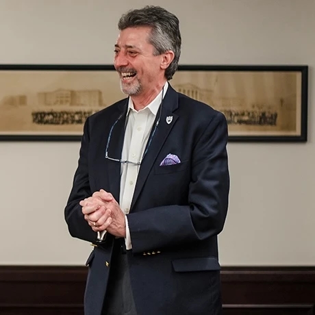 A smiling man in a suit with glasses hanging from his collar, standing in front of a framed picture.