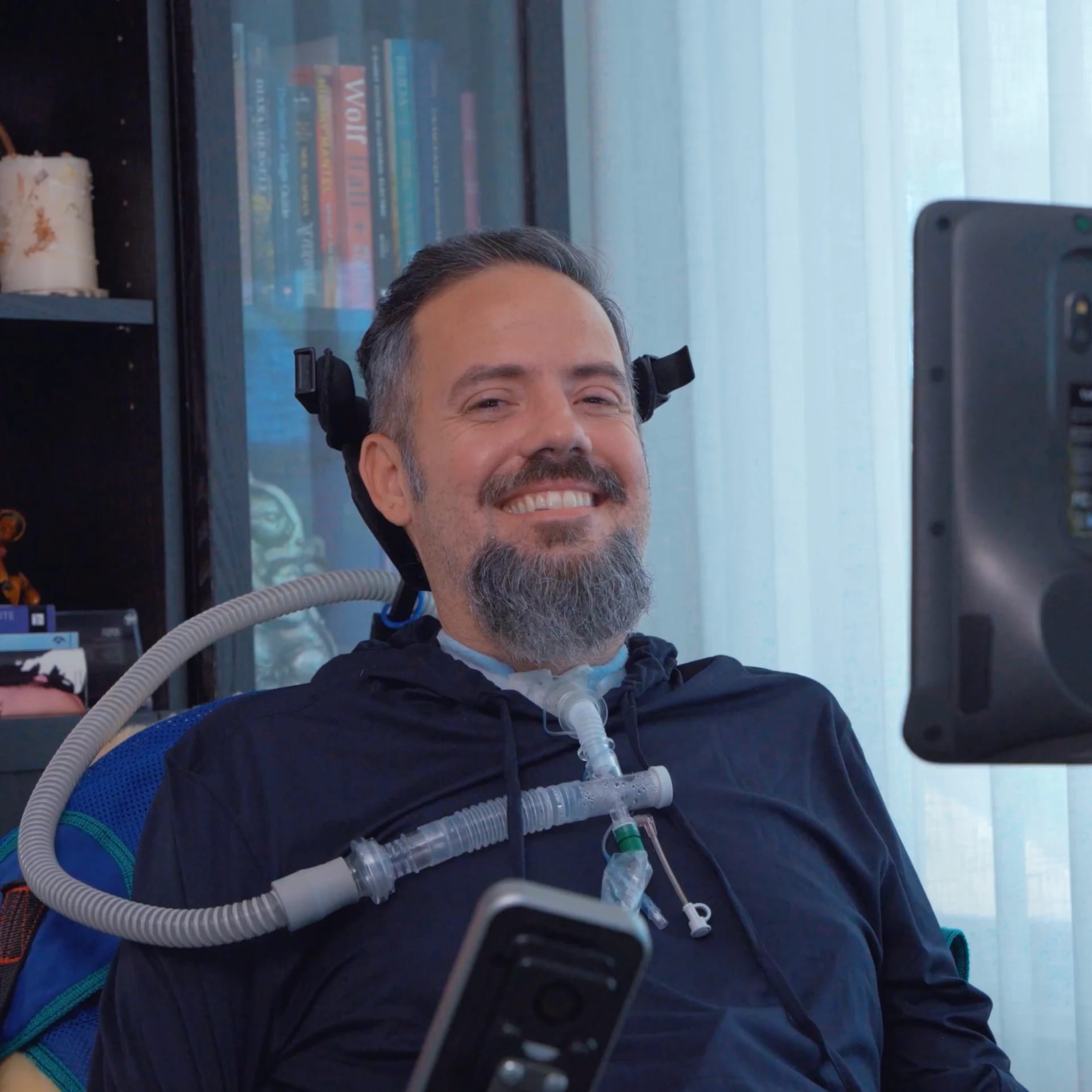 A man with a beard smiling, sitting in a wheelchair with medical equipment, in front of a bookshelf and a window.