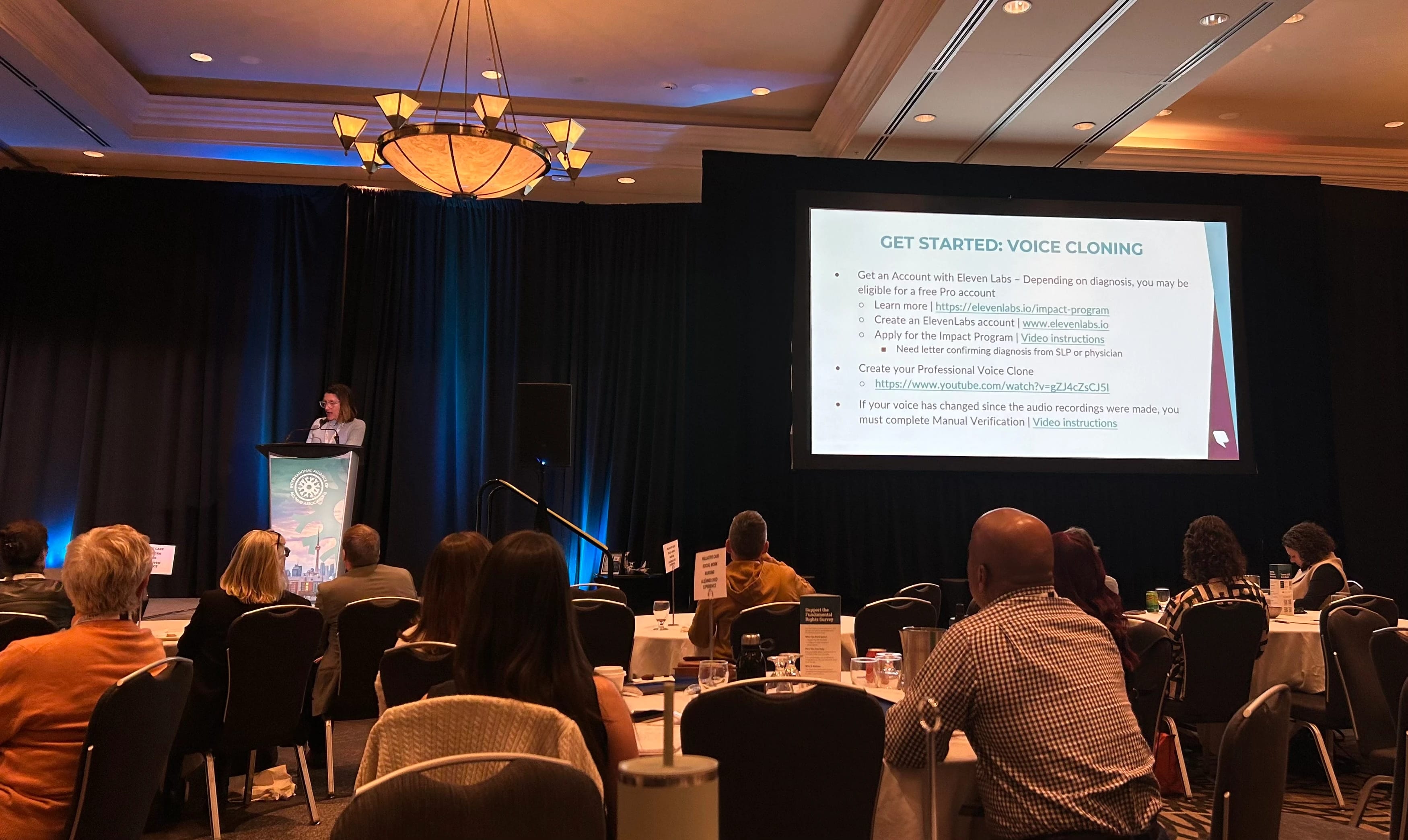 A woman standing at the podium at a conference in a hotel ballroom. Screen shows "Get Started: Voice Cloning"