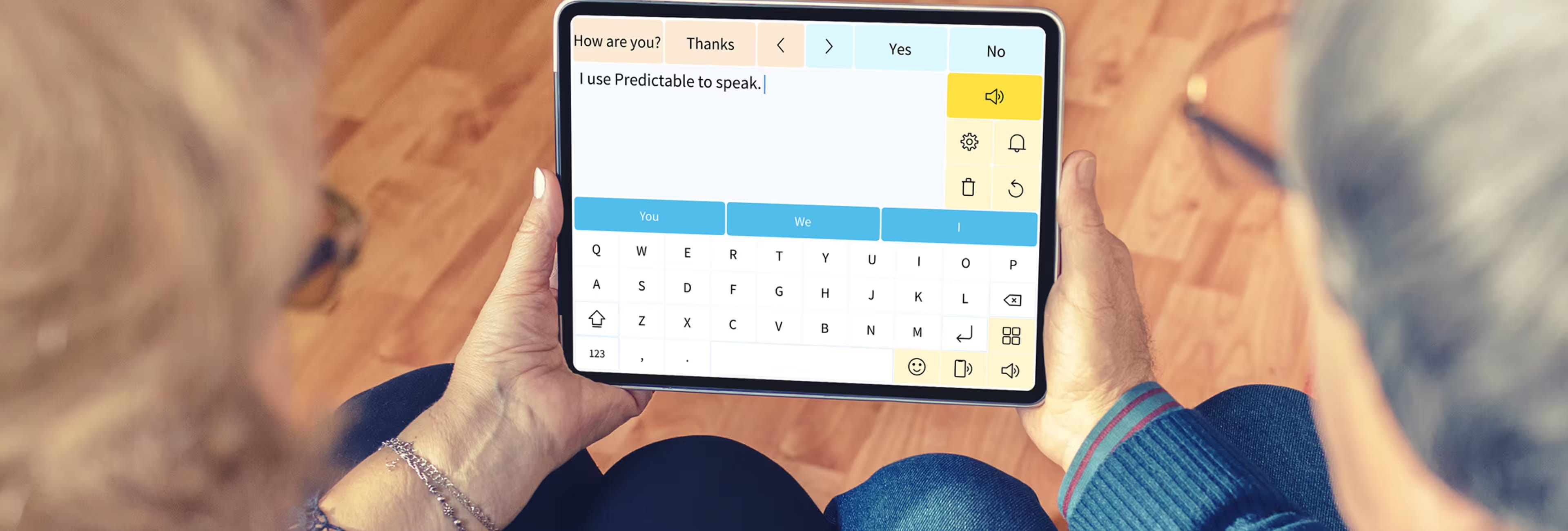 A person's hands are holding a tablet with the app "Predictable" open. The app's keyboard and a text field with the words "How are you? Thanks" and "I use Predictable to speak" are visible. The person is using the app to communicate.