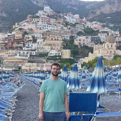 A man standing on a beach with rows of blue umbrellas and a hillside town in the background.