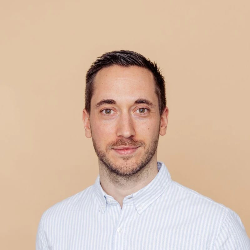 A man with short dark hair, light facial hair, wearing a light blue and white striped button-up shirt, smiling softly against a beige background.