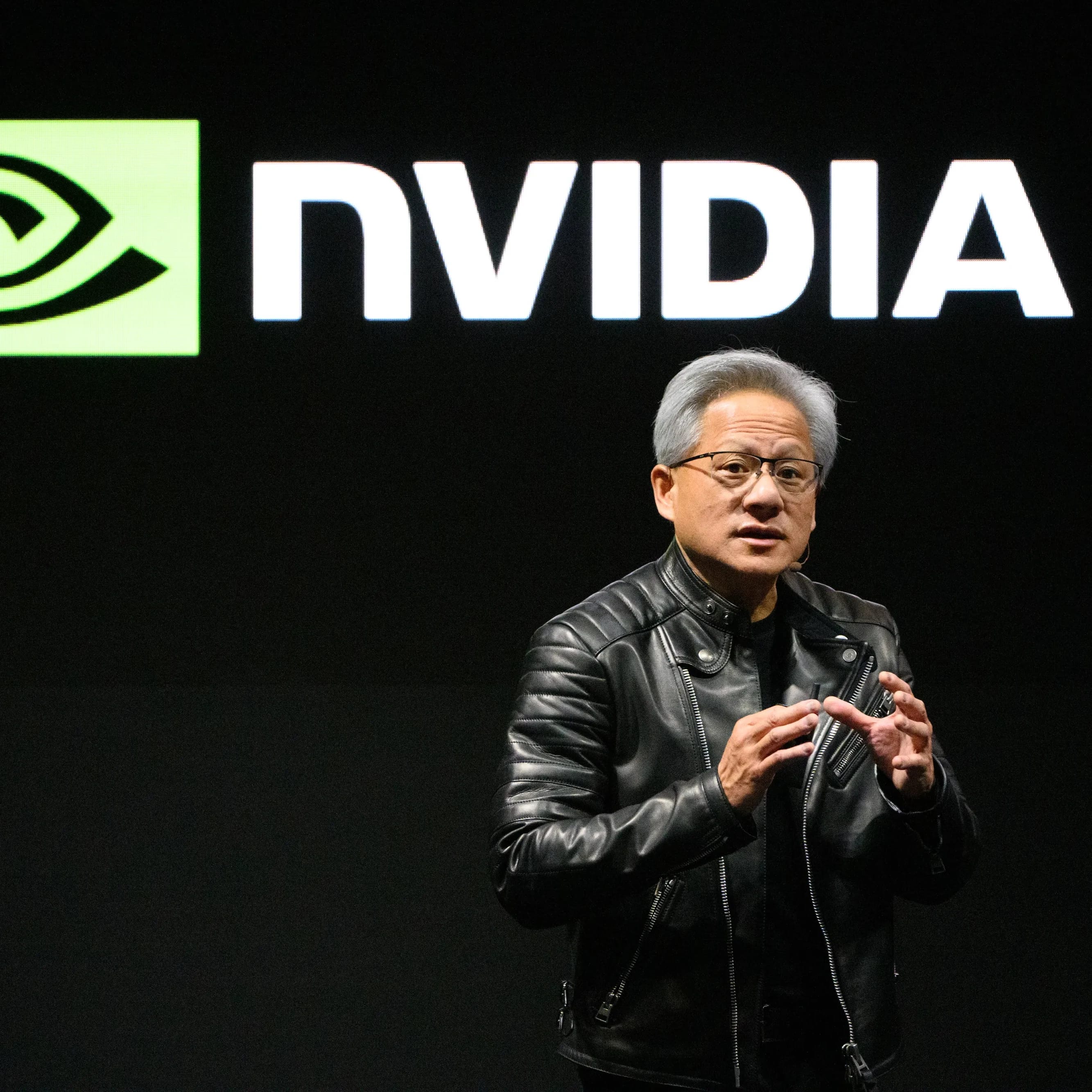 A man in a black leather jacket speaking in front of a large NVIDIA logo.