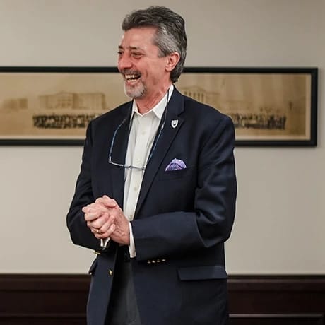 A smiling man in a suit with glasses hanging from his collar, standing in front of a framed picture.