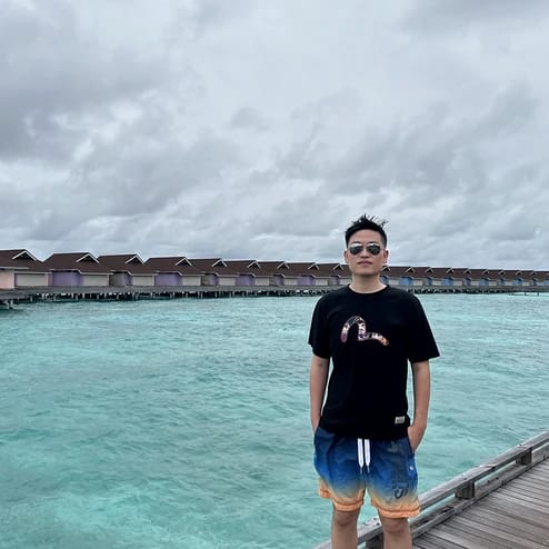 A young man wearing sunglasses and a black t-shirt with a snake graphic, standing on a wooden pier over clear turquoise water with overwater bungalows in the background.