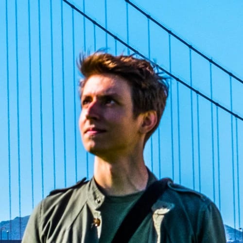 A young man standing outdoors in front of a blue bridge, looking into the distance.
