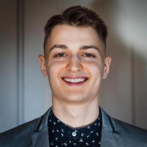 A young man with short brown hair, smiling, wearing a dark patterned shirt and a blazer.