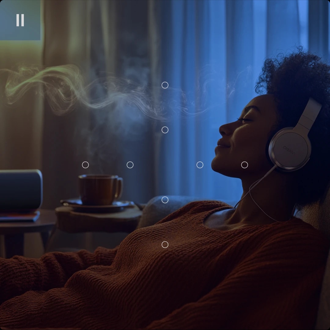 A woman relaxing with headphones on, smiling with eyes closed, in a cozy room with a steaming cup nearby.