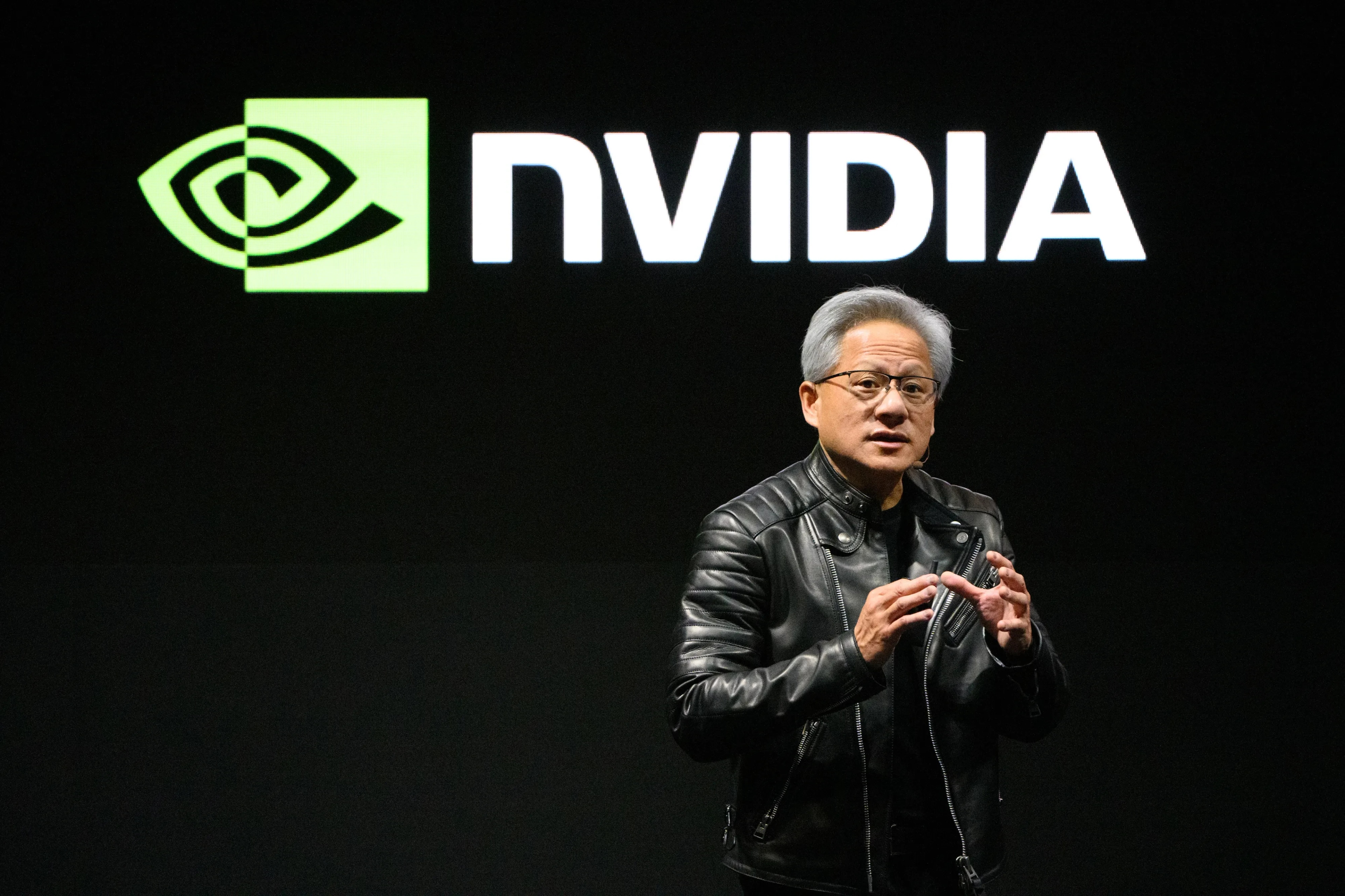 A man in a black leather jacket speaking in front of a large NVIDIA logo on a black background.