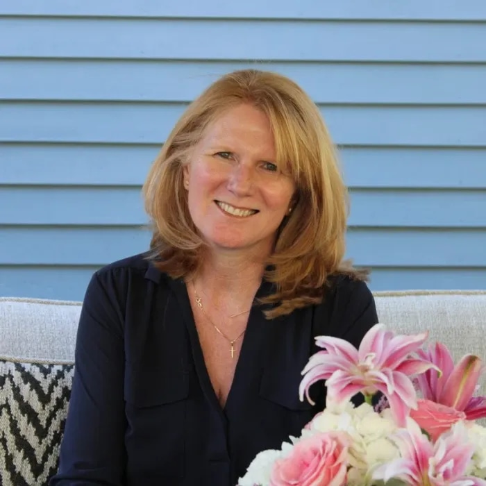 A woman with shoulder-length red hair smiling, sitting outdoors with a blue wooden wall in the background and a bouquet of pink and white flowers in front of her.