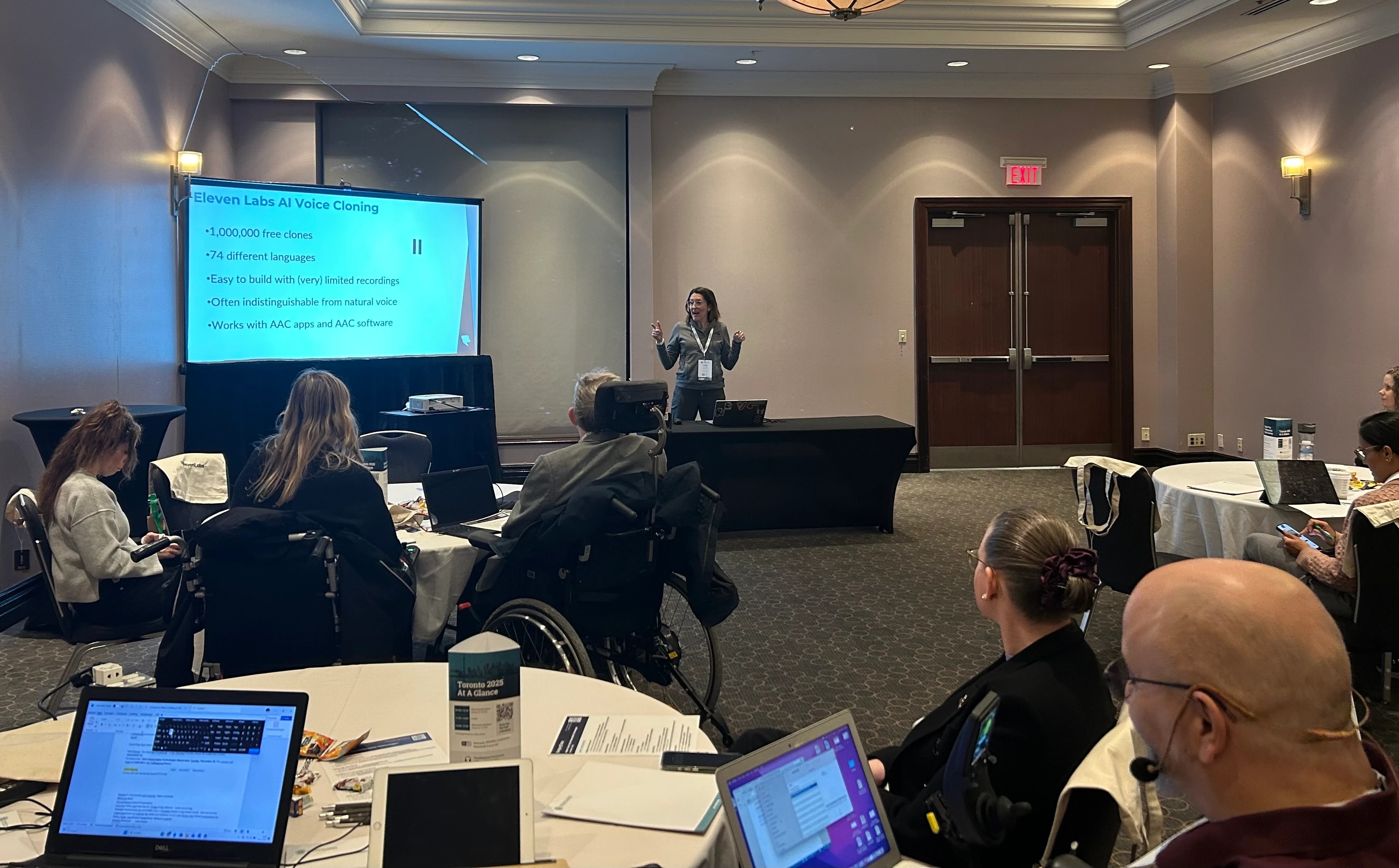 Woman standing in front of individuals sitting at tables, teaching a course. The screen behind her shows "ElevenLabs AI Voice Cloning"