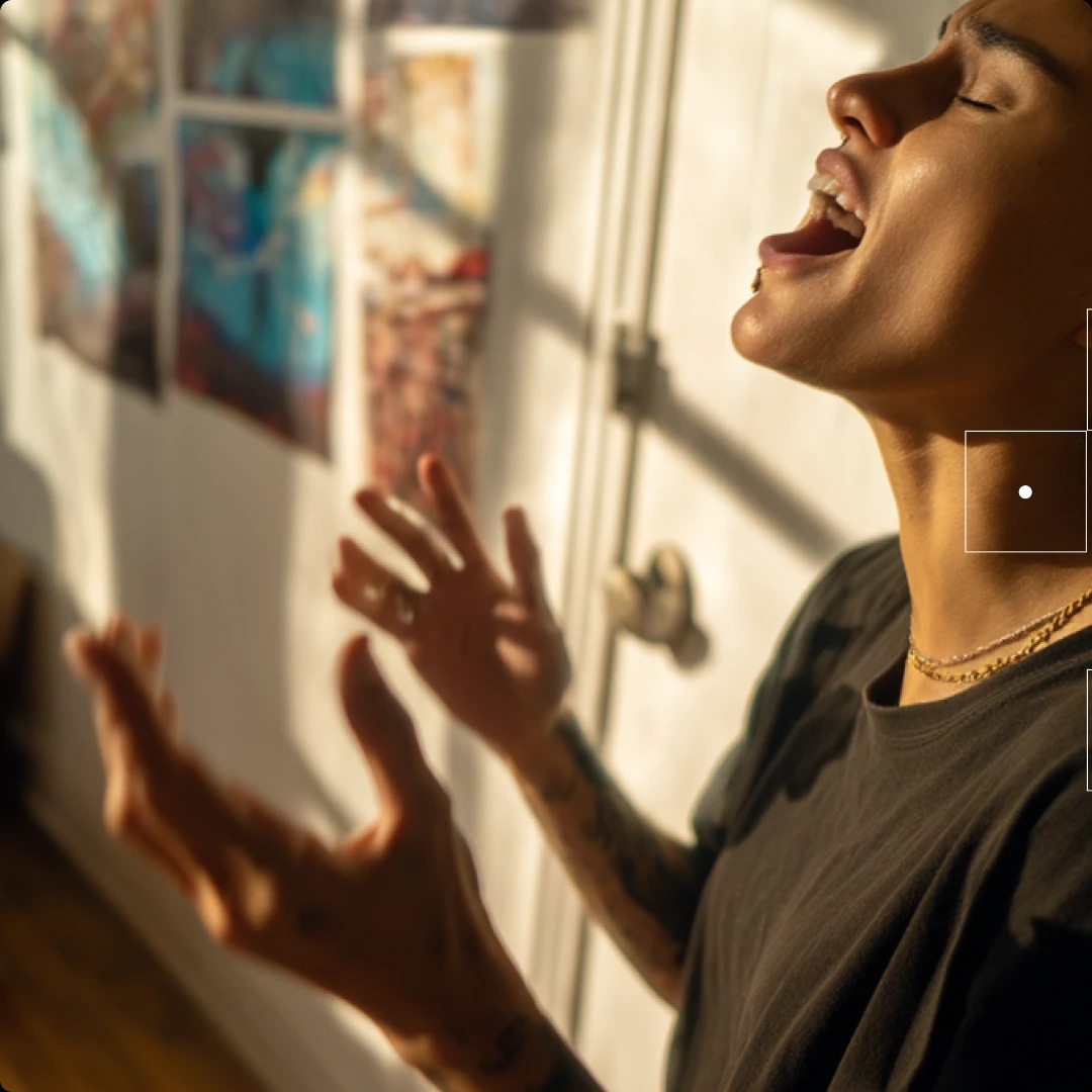 A person singing passionately with eyes closed, standing near a window with sunlight, and artwork on the wall behind.