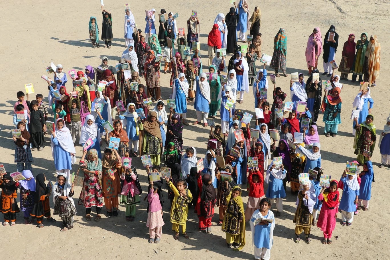 Group of children and women holding books outdoors on a sandy ground.