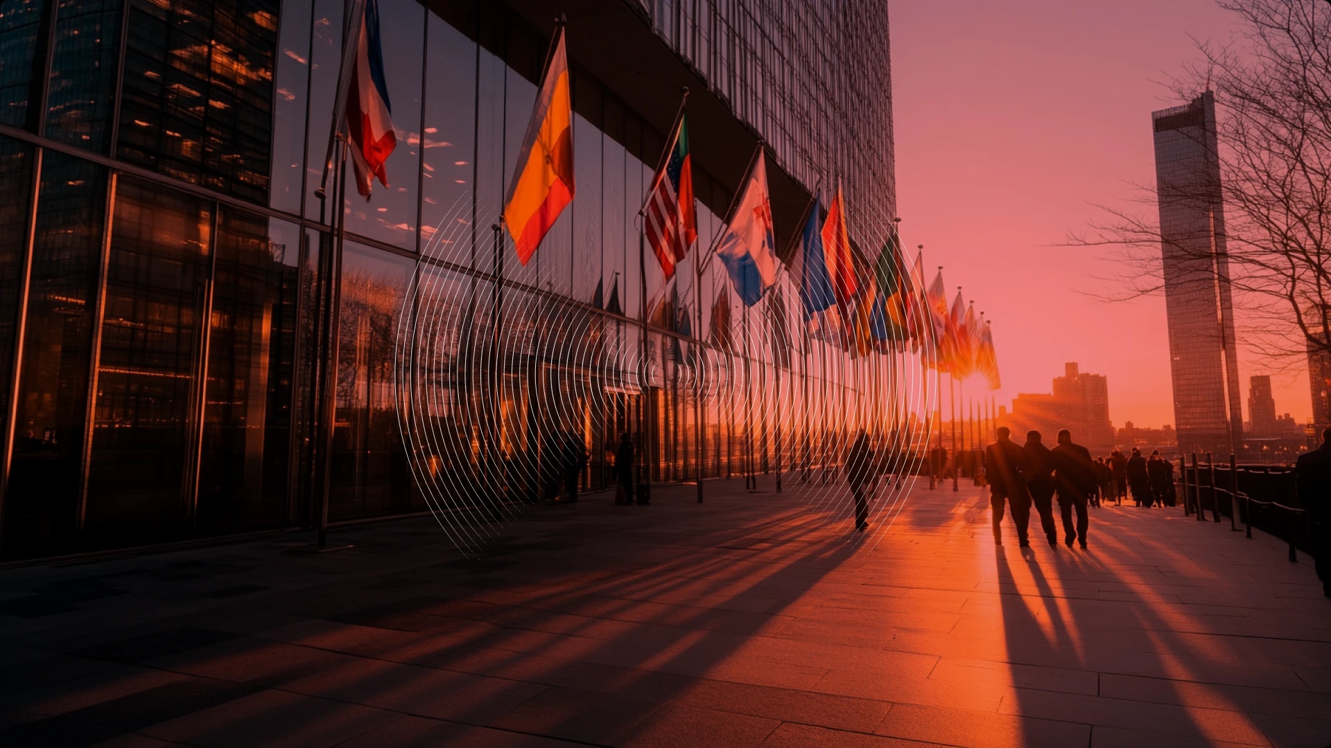 People walking outside a modern building with flags during sunset.