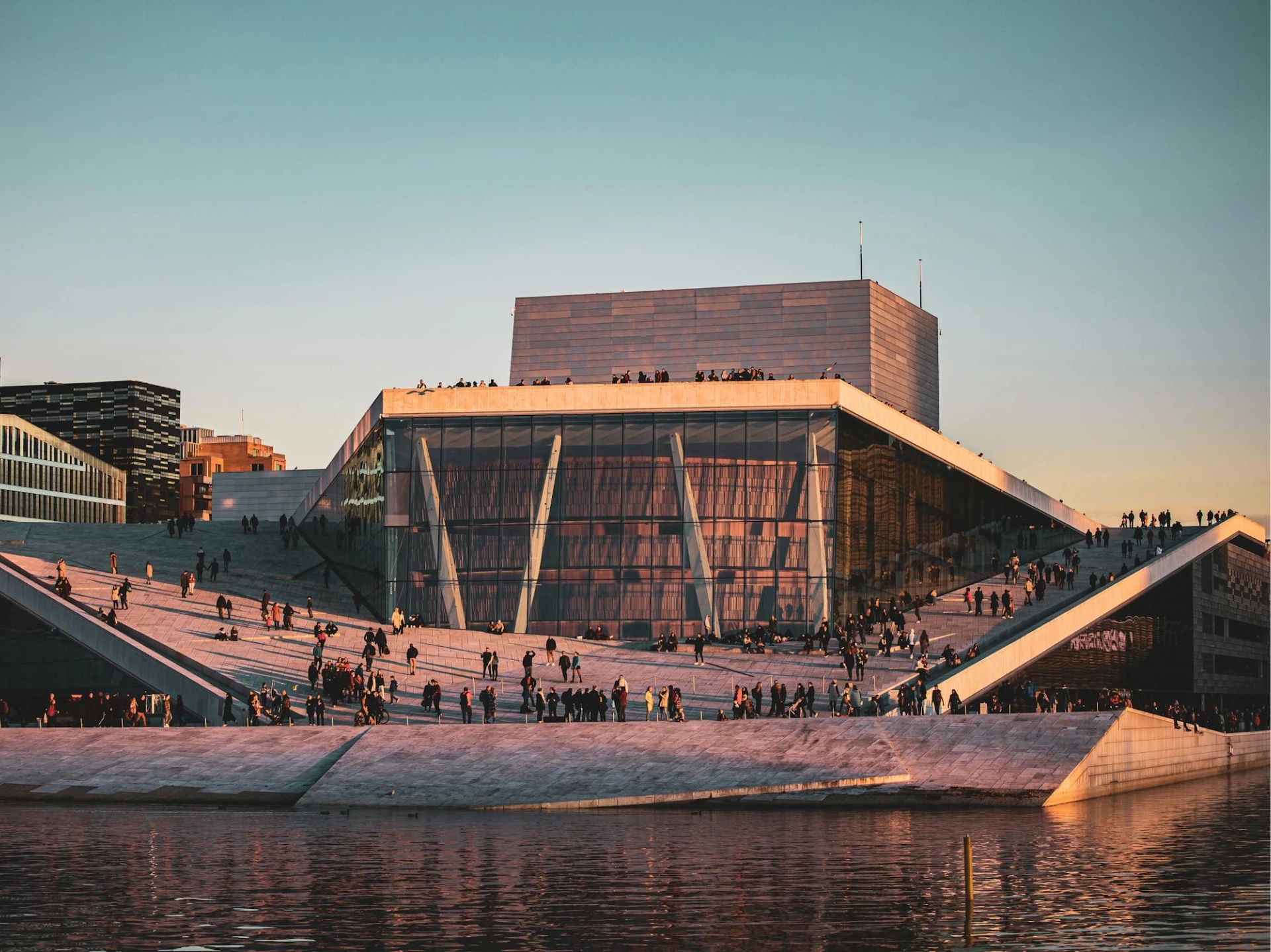 Modern building with glass facade and sloped walkways, filled with people, near a body of water at sunset.