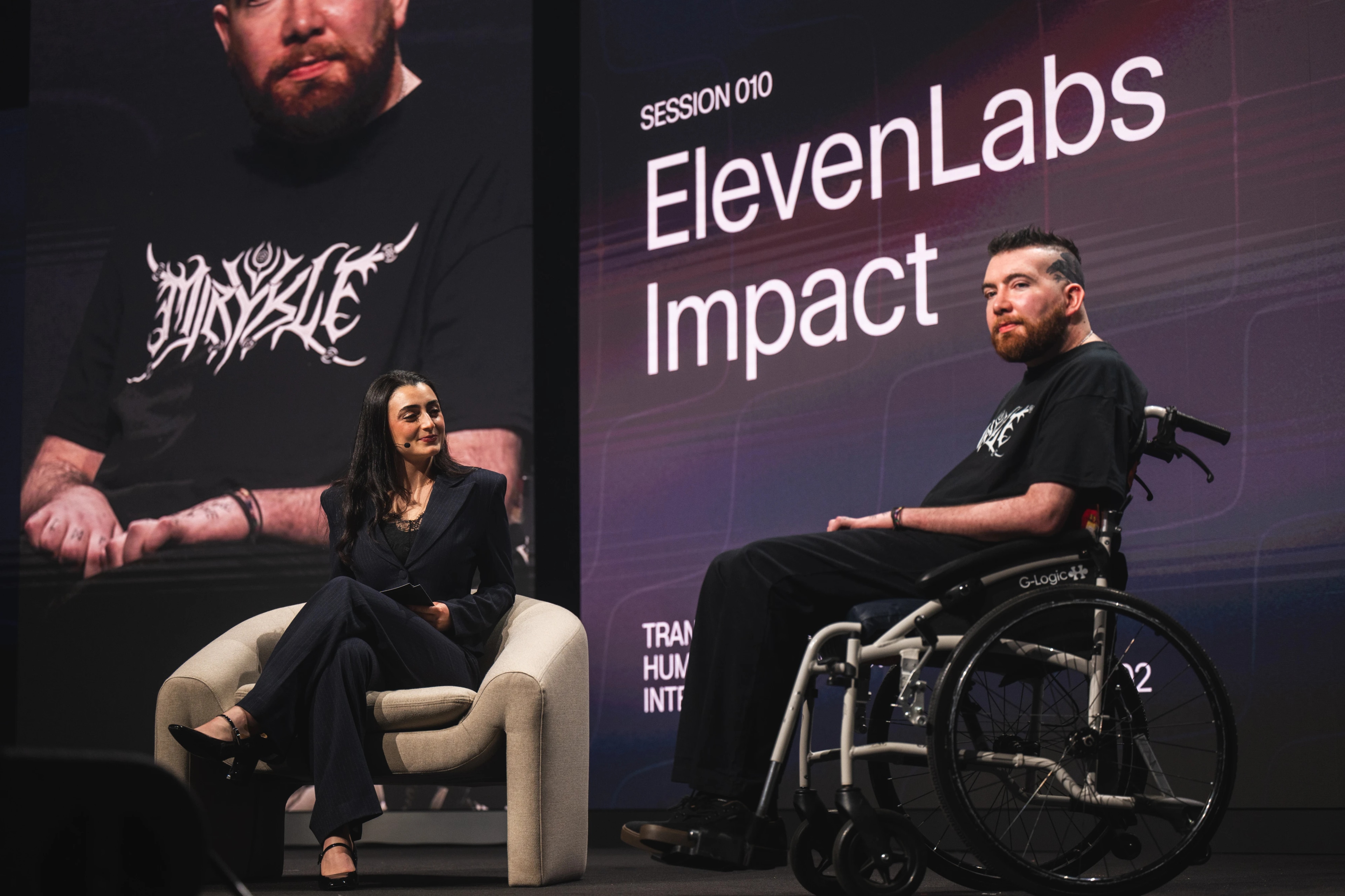 Man in a wheelchair sits on a stage across from a woman sitting in a chair, behind them is a large screen that reads "ElevenLabs Impact"
