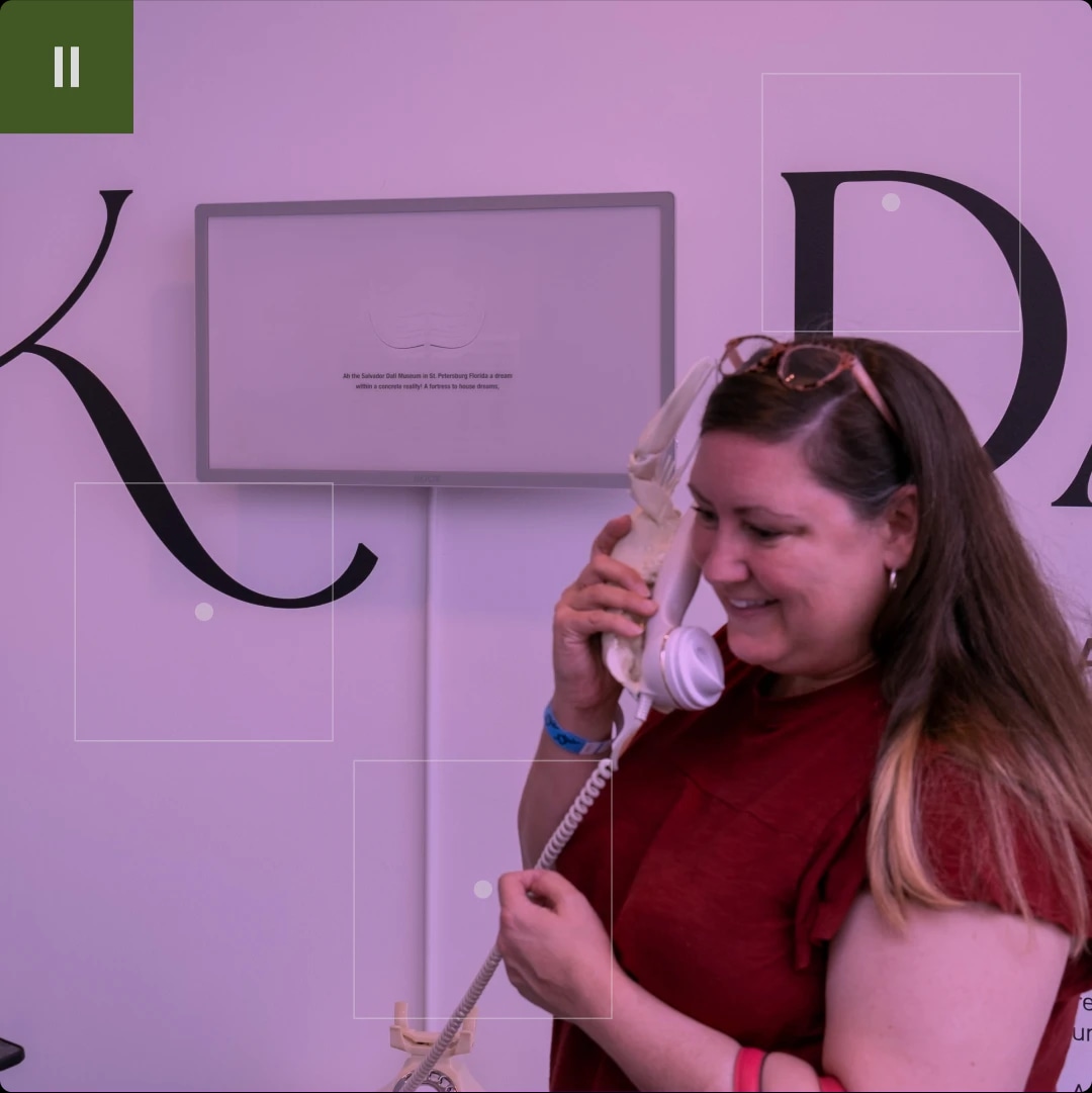 A woman smiling while talking on a vintage telephone with a skull-shaped base, in front of a display with large black letters on a purple wall.