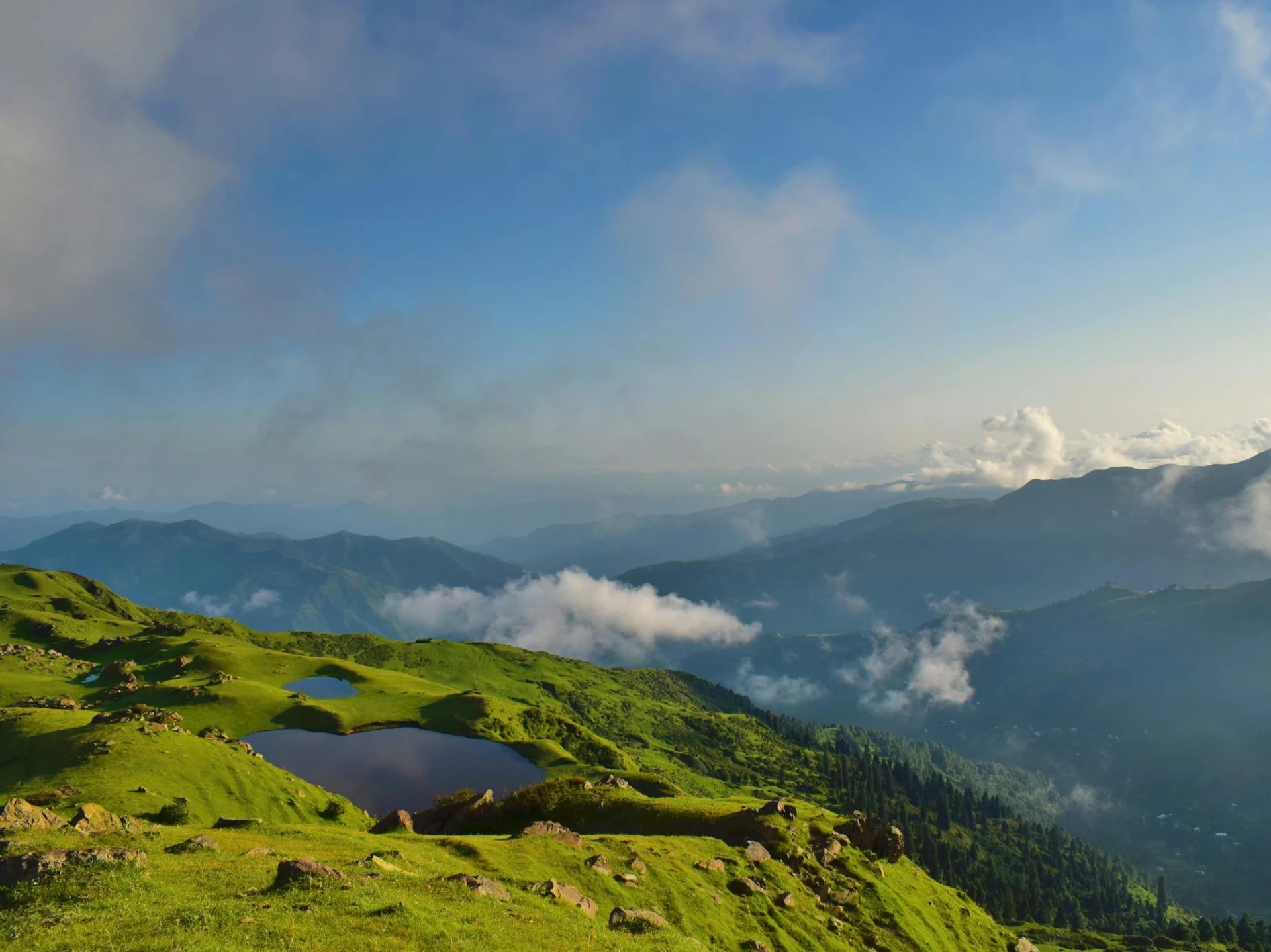 Green rolling hills with small lakes, mountains in the background, and a partly cloudy sky.