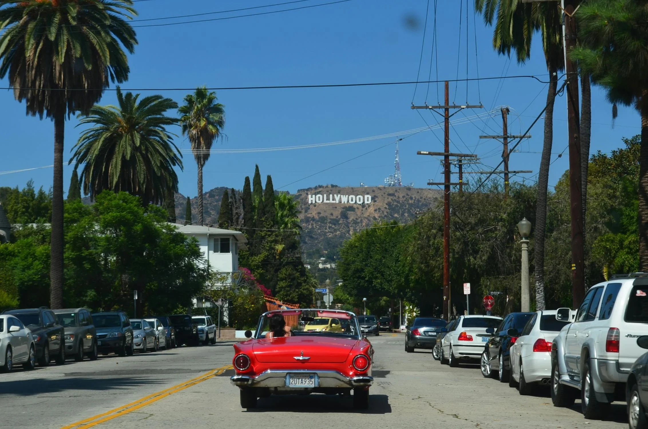 A red vintage car driving down a street with the Hollywood sign visible on the hillside in the background.