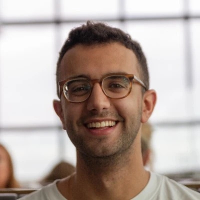 A smiling man with glasses in an indoor setting with large windows in the background.