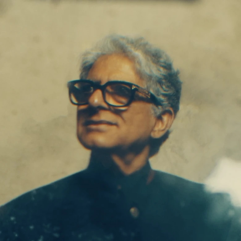 A person with gray hair wearing large black glasses and a dark high-collared shirt, looking slightly to the side.