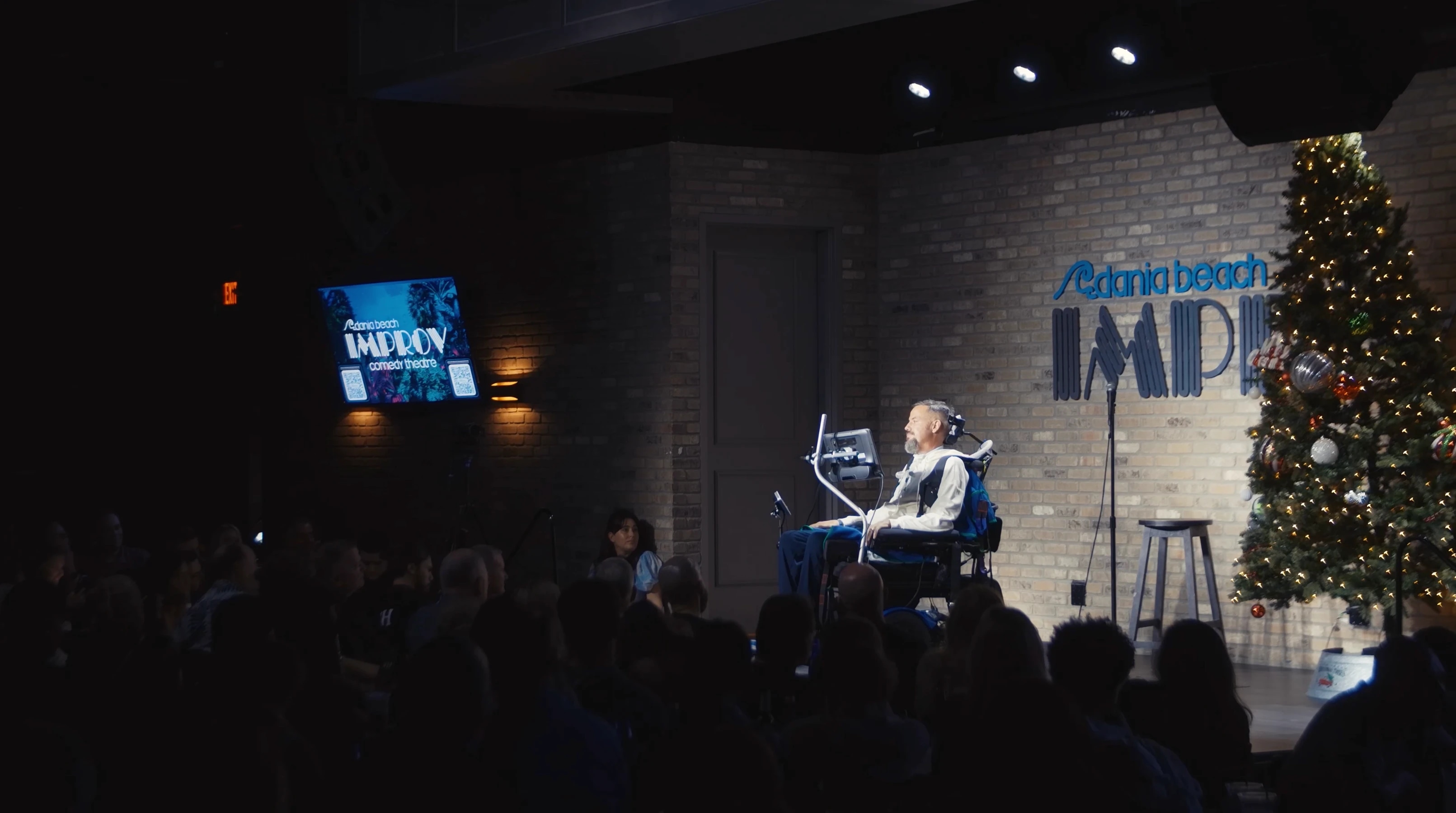 A man in a wheelchair performing on stage at the Improv comedy theater, decorated for Christmas with a Christmas tree.