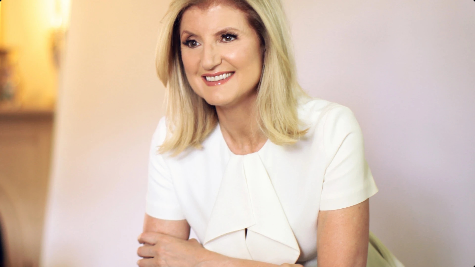A woman with blonde hair smiling, wearing a white top, sitting with her arms crossed.