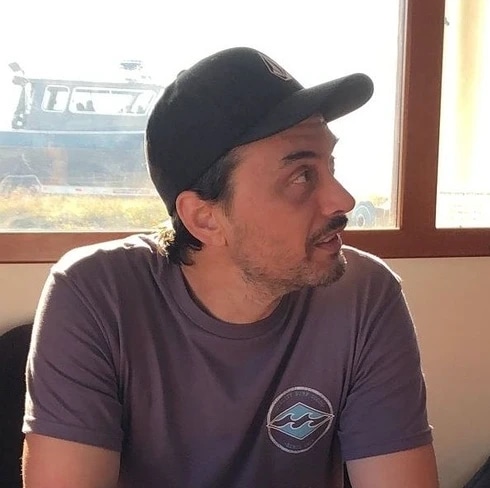 A man wearing a black cap and a gray t-shirt sitting indoors near a window, looking to his right.