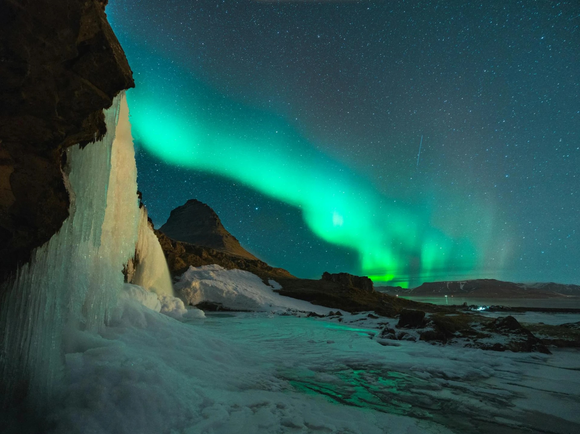 Northern lights over a snowy landscape with a waterfall and mountain in the background.