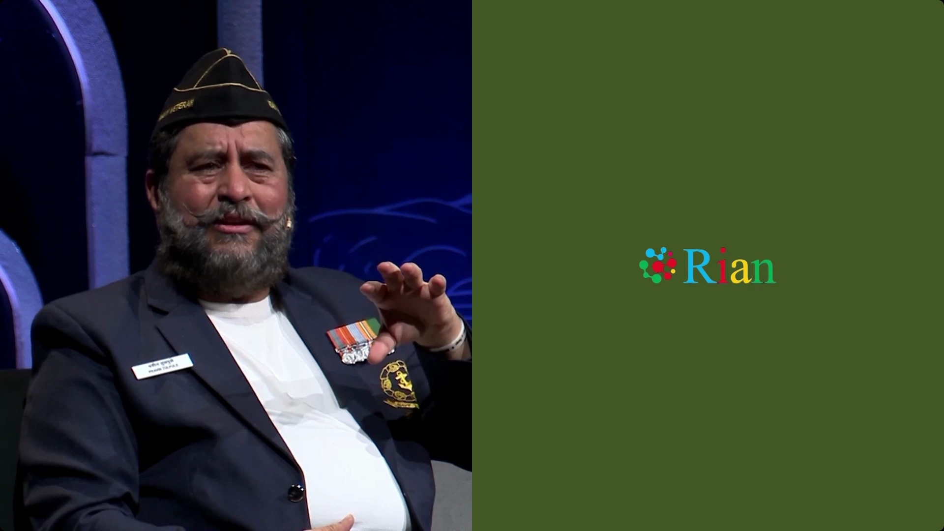 A man with a beard and mustache, wearing a military-style hat and a suit with medals, sitting and gesturing with his hand.