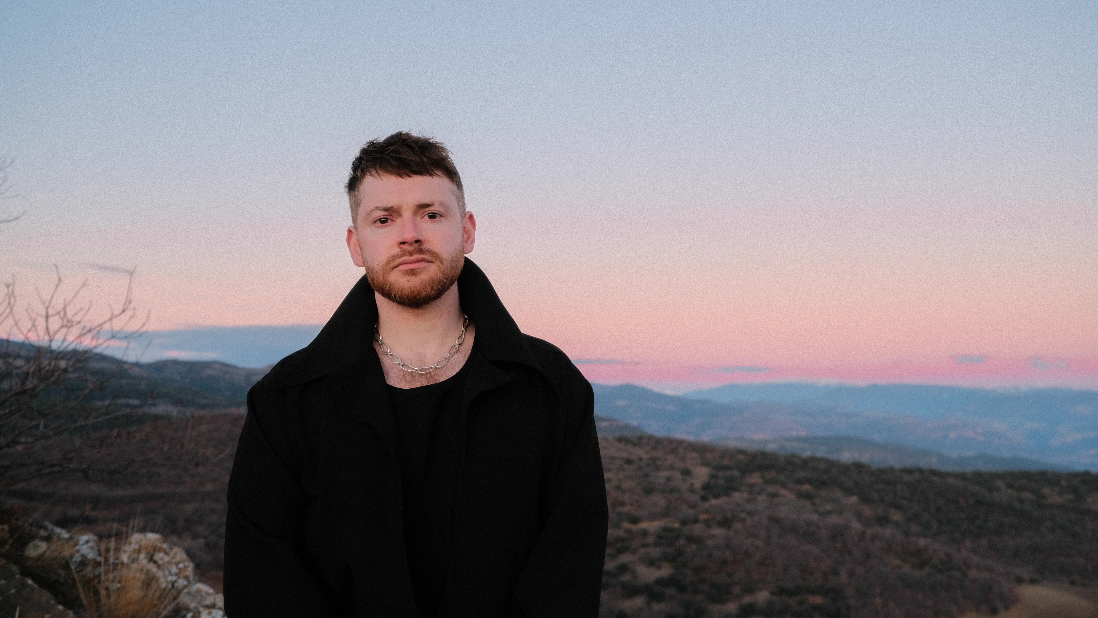 A man with short hair and a beard standing outdoors during sunset, wearing a black jacket and a silver chain necklace, with a landscape of mountains and a pink and blue sky in the background.