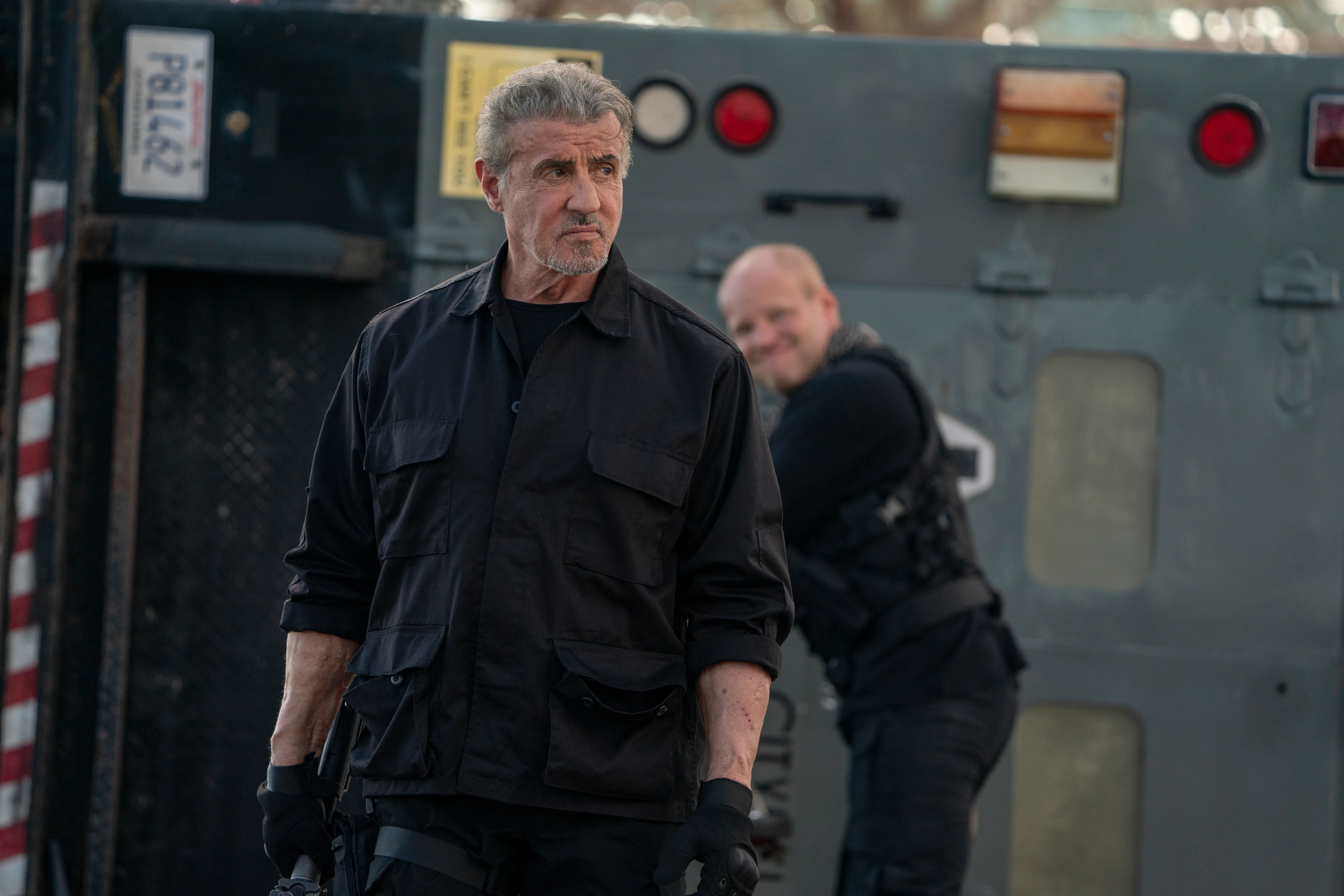 A man with gray hair and a beard, dressed in black tactical gear, stands in front of a large vehicle, with another man smiling in the background.