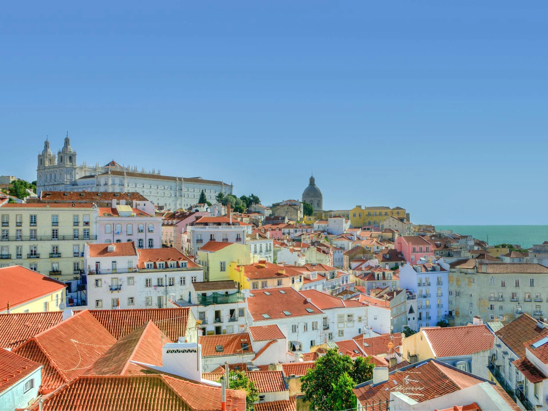 A panoramic view of a coastal city with colorful buildings and red-tiled roofs, featuring a large church and a domed structure on a hill, with the ocean in the background.