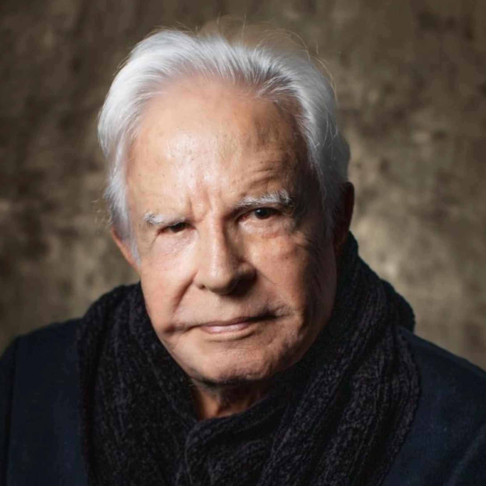 An elderly man with white hair, wearing a dark scarf, looking directly at the camera with a serious expression.