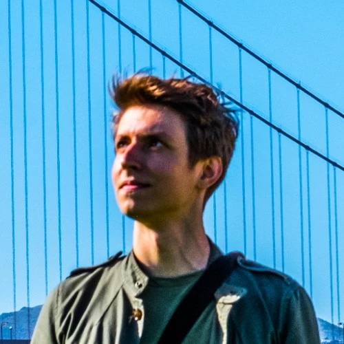 A young man standing outdoors in front of a blue bridge, looking into the distance.