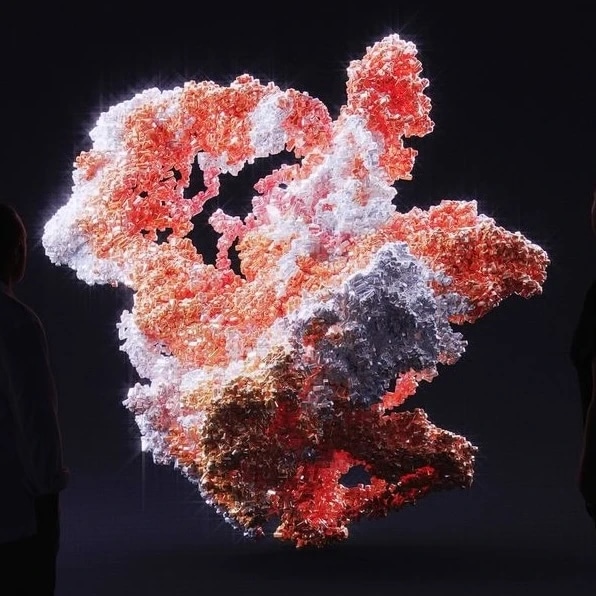 A large, abstract, illuminated sculpture with pink, red, white, and black colors, viewed against a dark background with two people observing it.