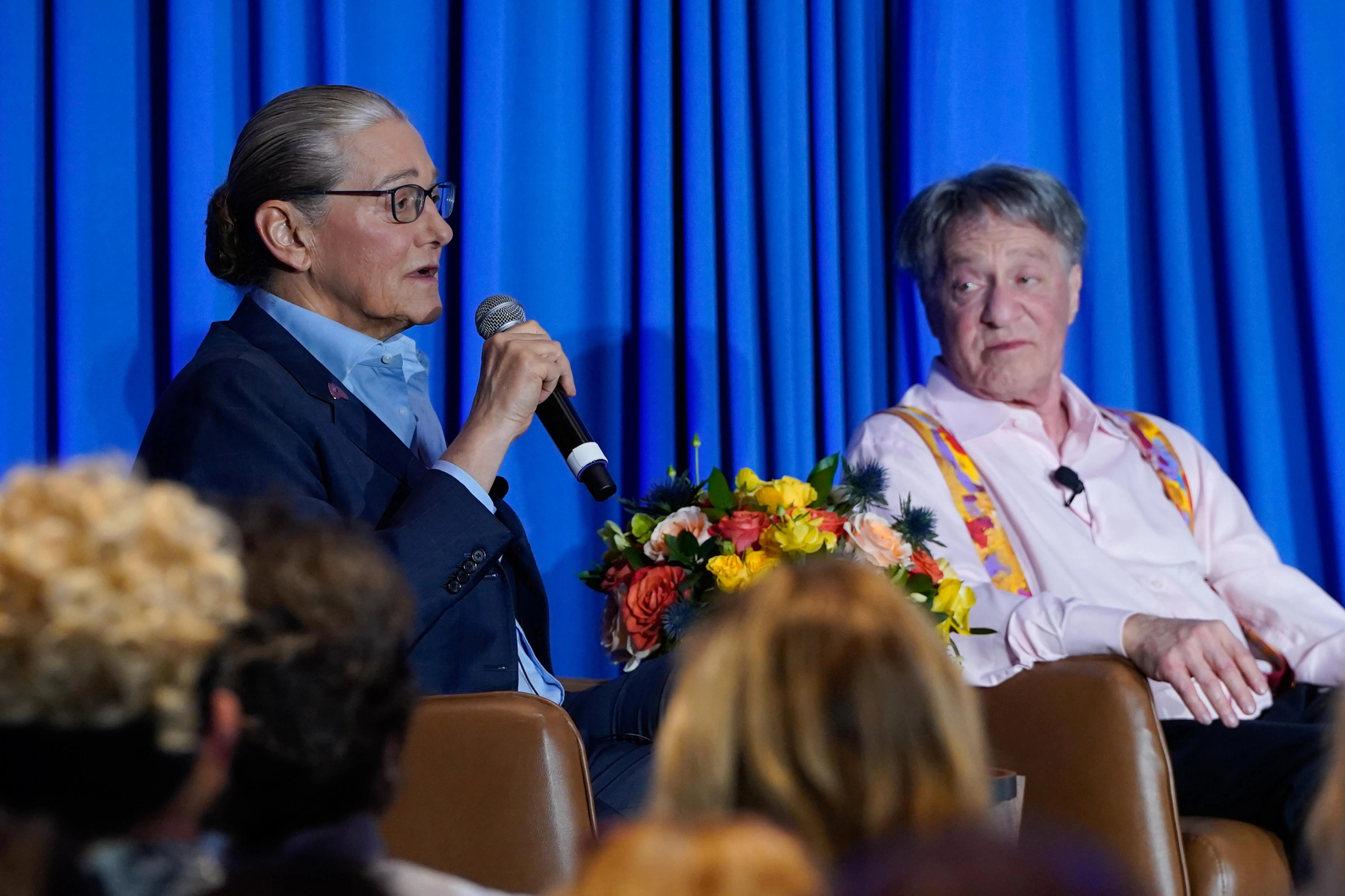 Two people onstage speaking, one into a microphone in front of a blue backdrop