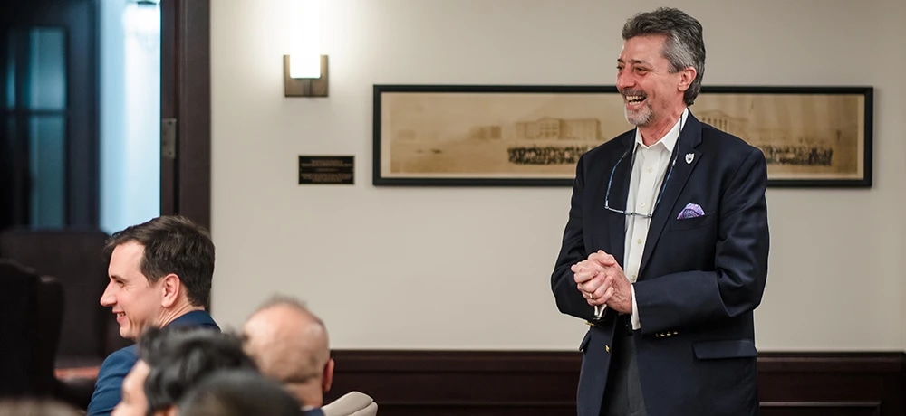 A man in a suit smiling and clapping in a room with an audience.