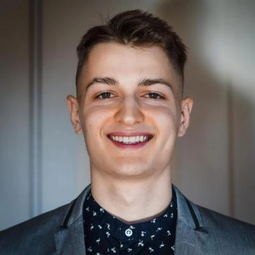 A young man with short brown hair, smiling, wearing a dark patterned shirt and a blazer.