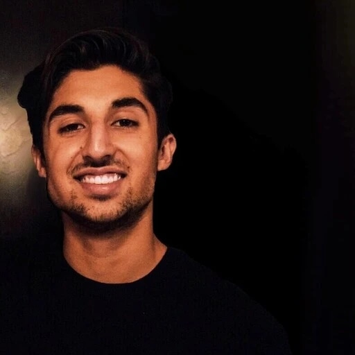 A young man with dark hair and a beard smiling at the camera against a dark background.