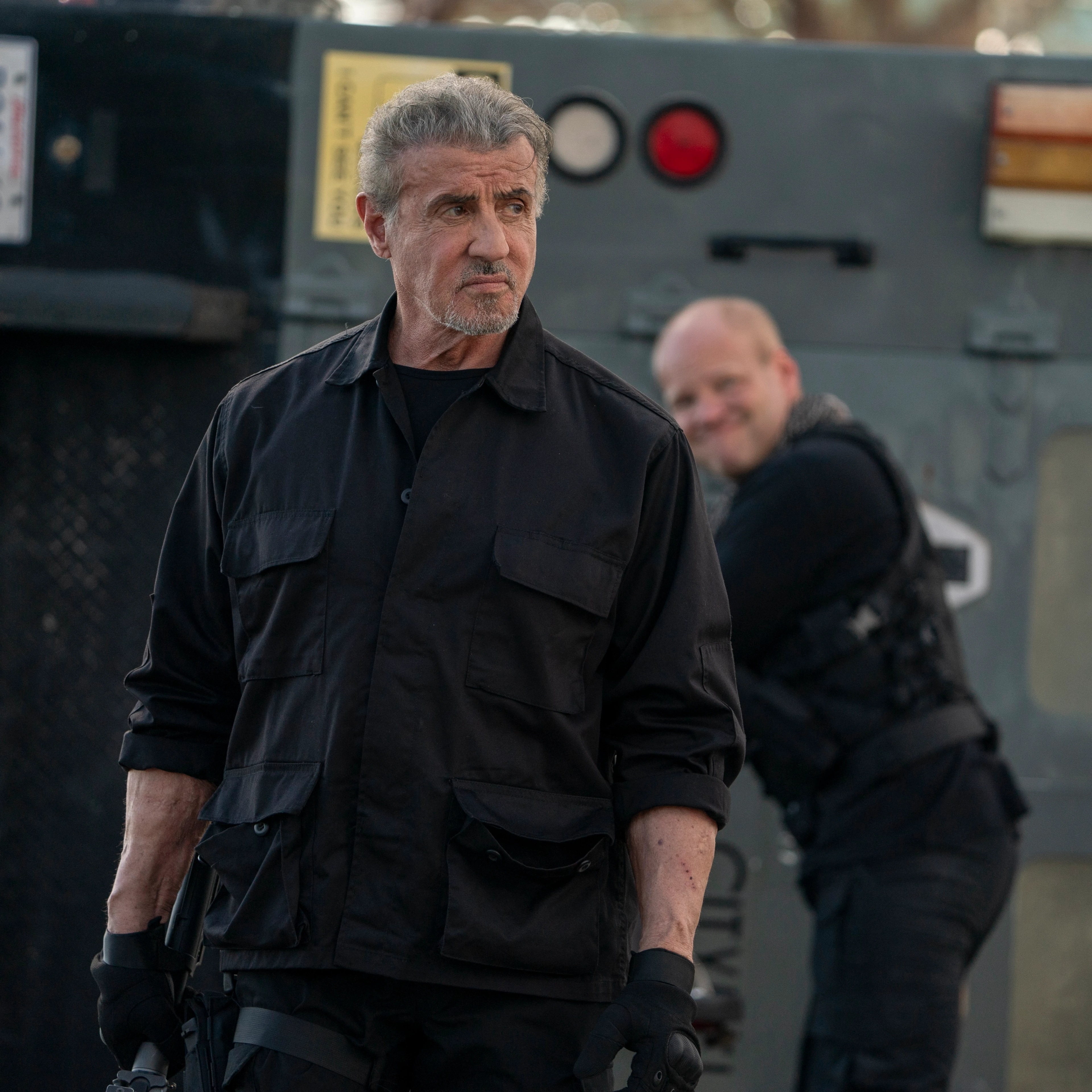 A man with gray hair and a beard, dressed in black tactical gear, holding a gun, standing outdoors with a serious expression.