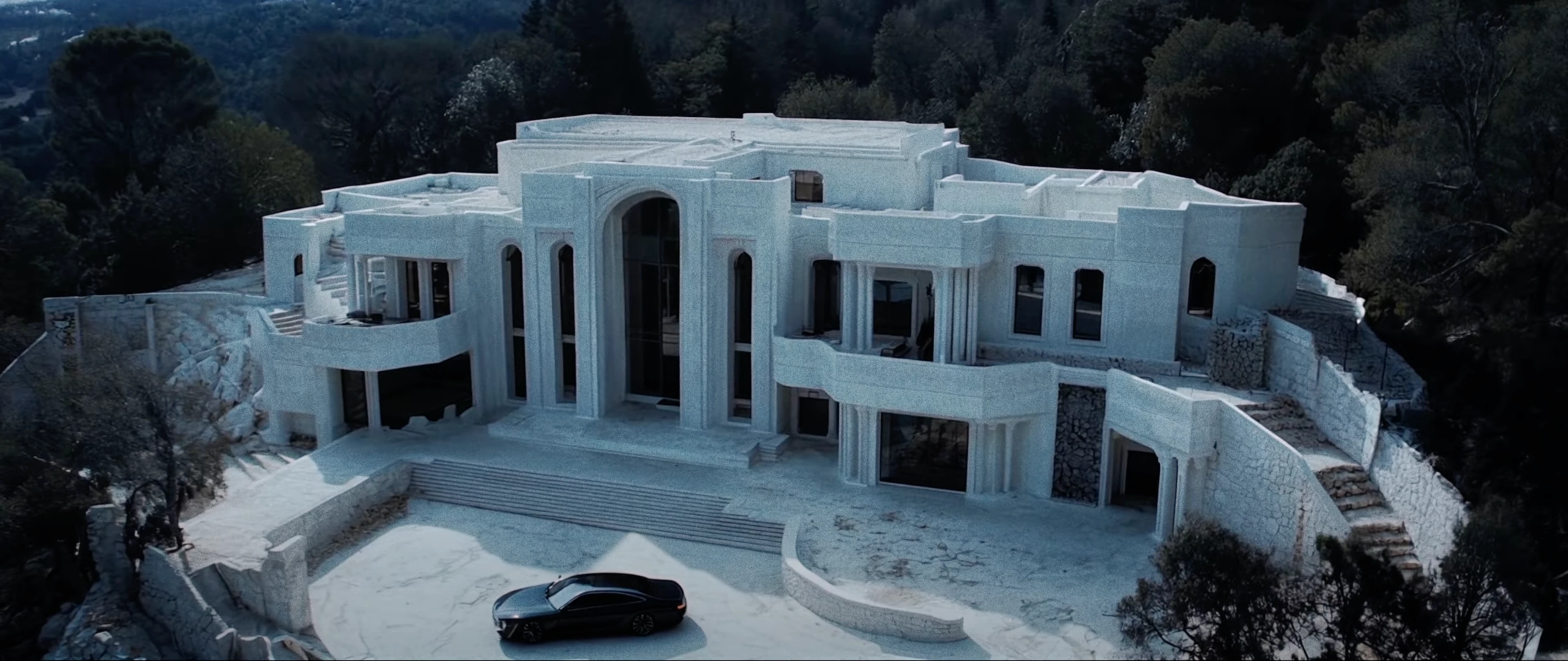 A large, modern white mansion on a hillside surrounded by trees, with a black car parked in front.