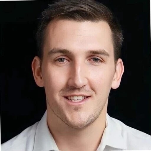 A young man with short brown hair, smiling, wearing a white shirt against a dark background.