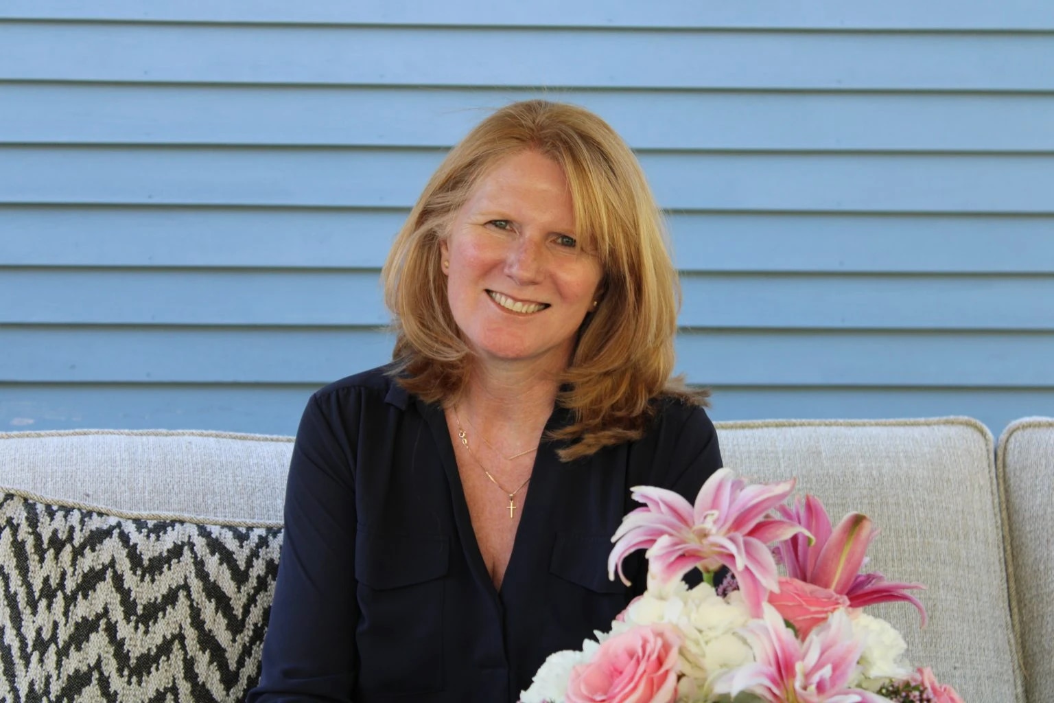 A woman with shoulder-length red hair smiling while sitting on a light-colored outdoor sofa with a bouquet of pink and white flowers.