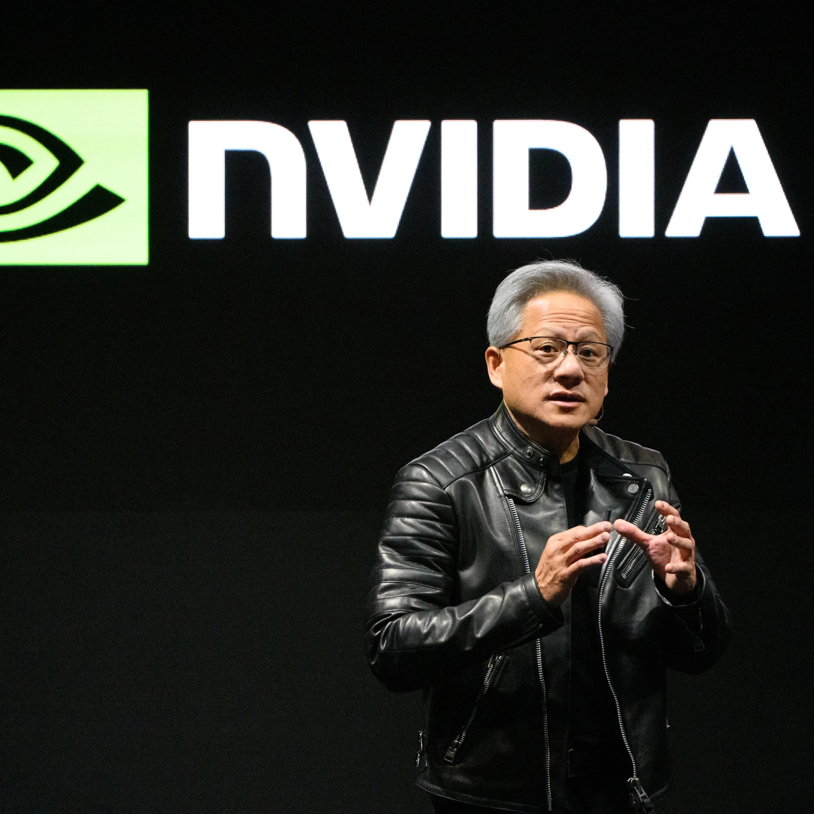 A man in a black leather jacket speaking in front of a large NVIDIA logo.