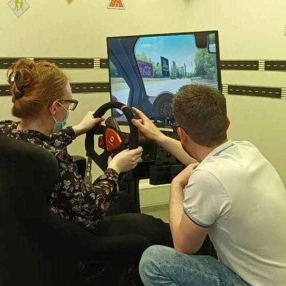 Two children, one girl and one boy, are sitting in front of a driving simulator screen. The girl is holding the steering wheel, and the boy is pointing at the screen.