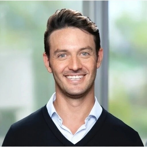 A smiling man with dark hair and a white shirt, standing in front of a window.