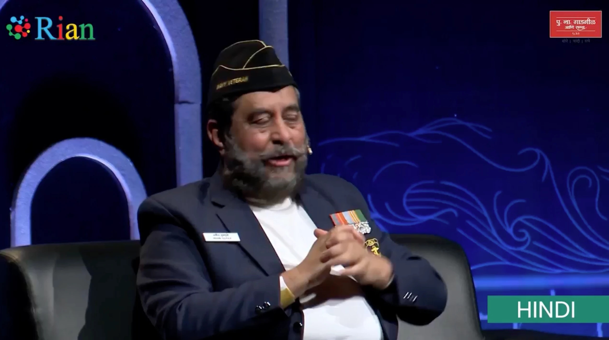 A man with a beard and mustache, wearing a black cap and a dark blazer with medals, is sitting on a black chair with his hands clasped.