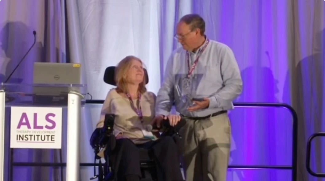 A woman in a wheelchair holding hands with a man on stage at an ALS Institute event.