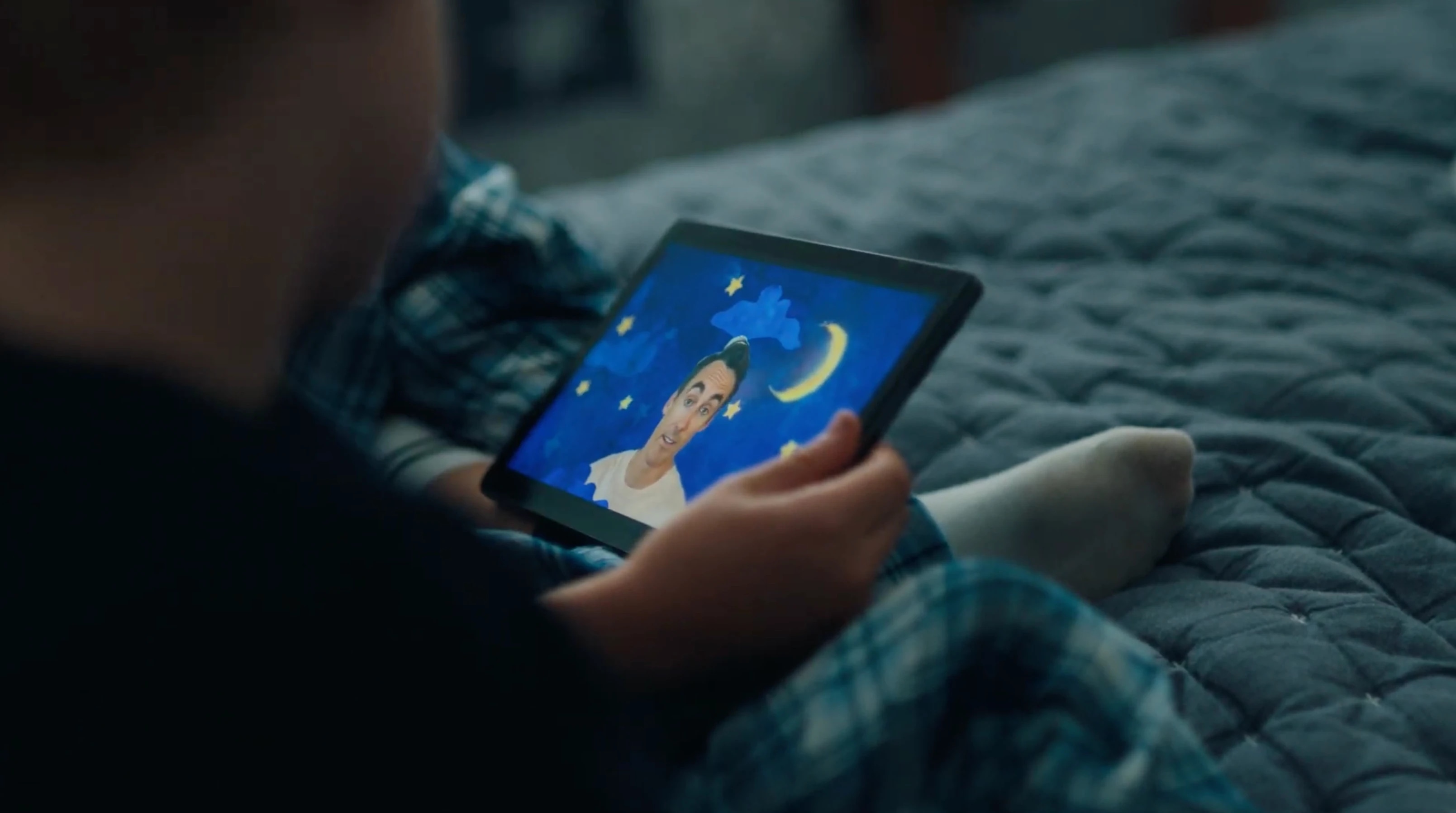 A person lying on a bed holding a tablet displaying a cartoon character with a night sky background.